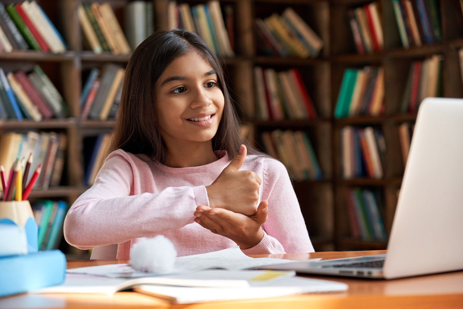 Smiling Indian girl with hearing disability in school library on a laptop communicating with teacher by video conference call using sign language showing hand gesture during virtual lesson.