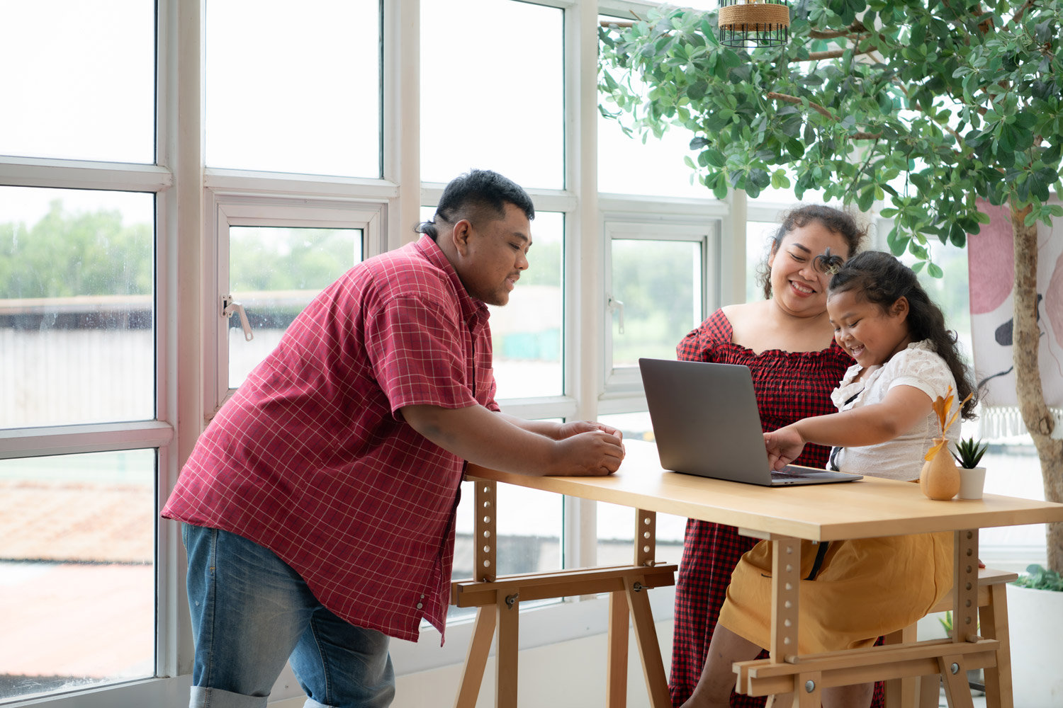 Hispanic family with a father wearing a prosthetic leg, is happily assisting a child as she uses a laptop. Behind them is a bright, office building with large windows.