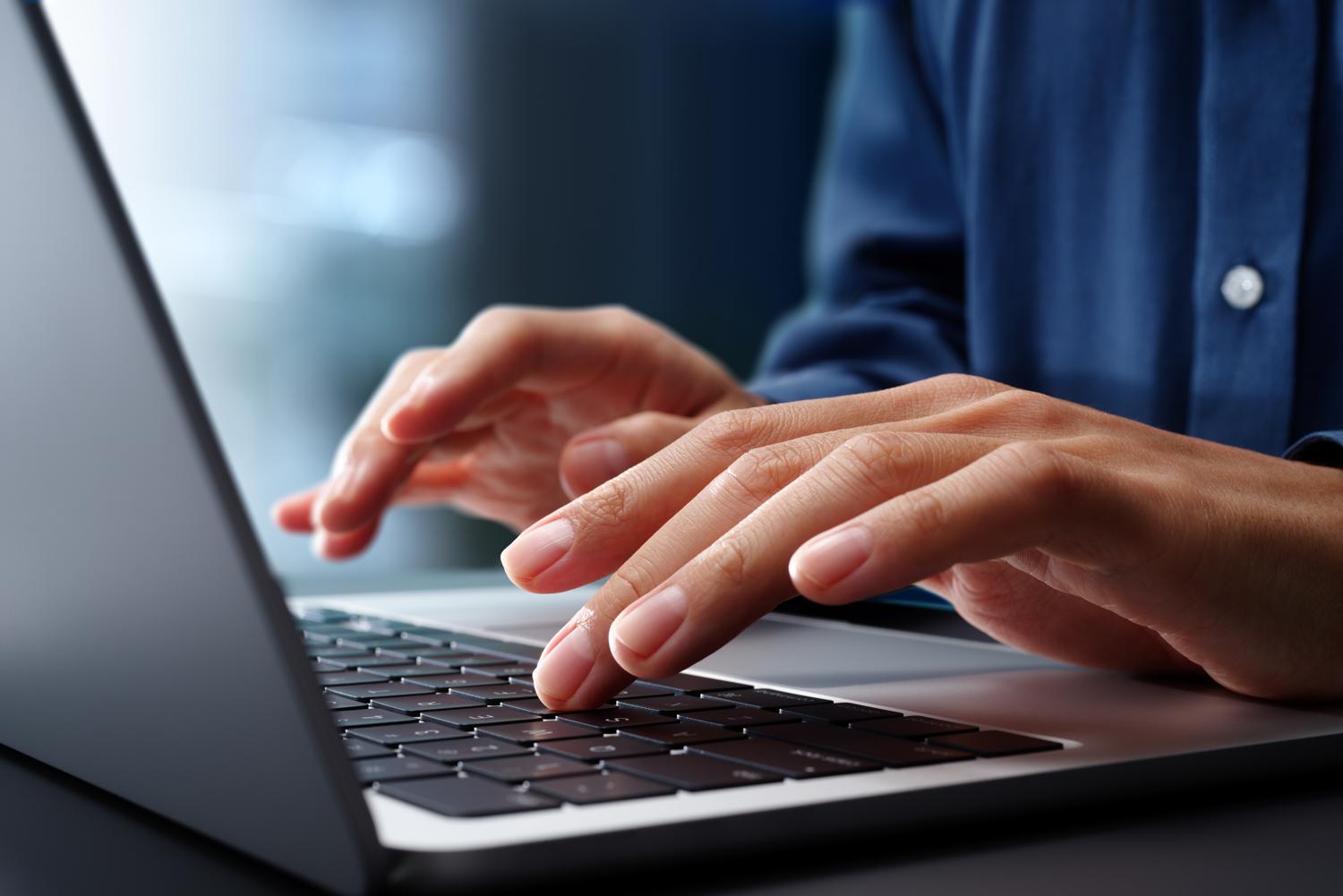 Close up of hands typing on laptop computer keyboard. Person is wearing a blue button-down shirt.