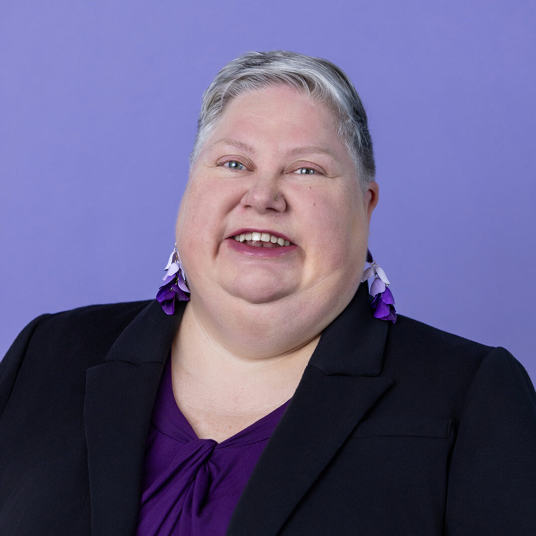 A person with short gray hair wearing a dark blazer and a purple top, stands against a light purple background. They are smiling, facing the camera and wearing purple and white earrings.
