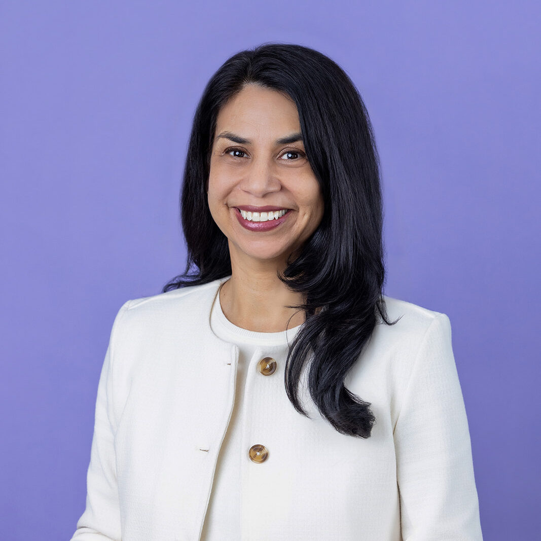 A person with long dark hair wearing a white jacket and top stands facing the camera against a light purple background.