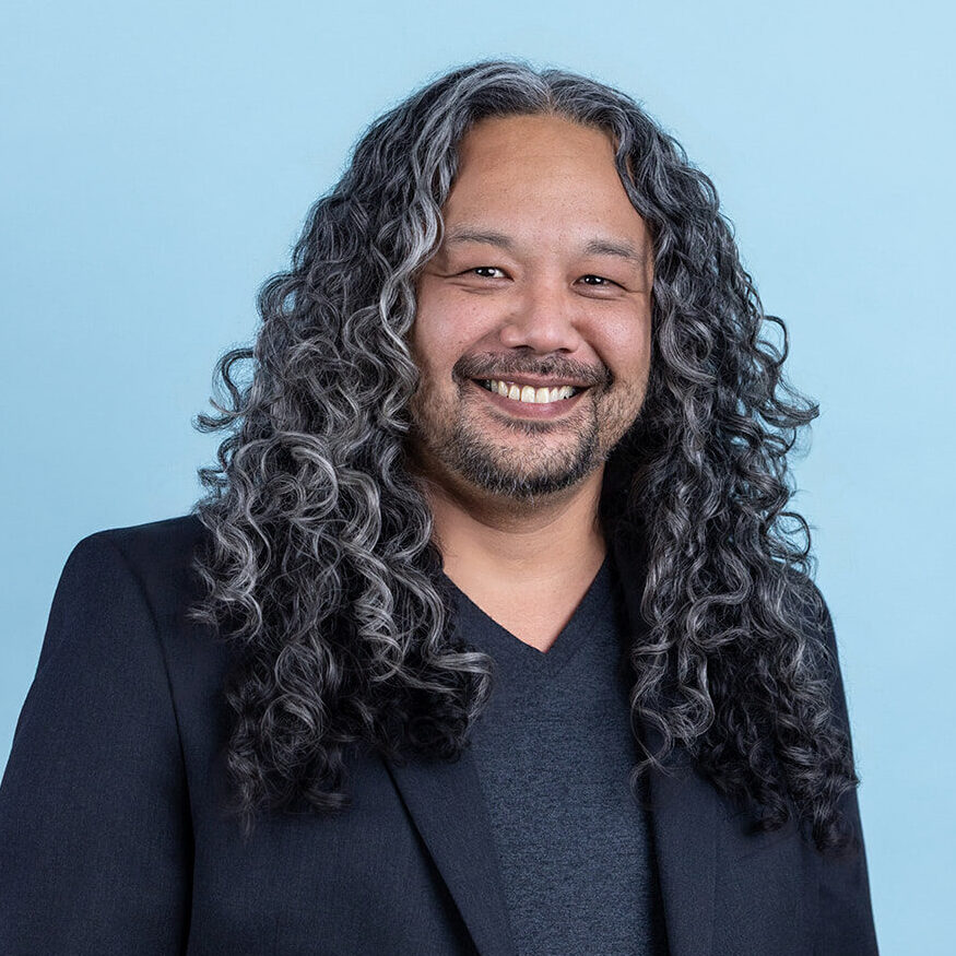 A person with long curly gray and black hair wearing a dark blazer over a dark V neck shirt stands in front of a light blue background, facing the camera.