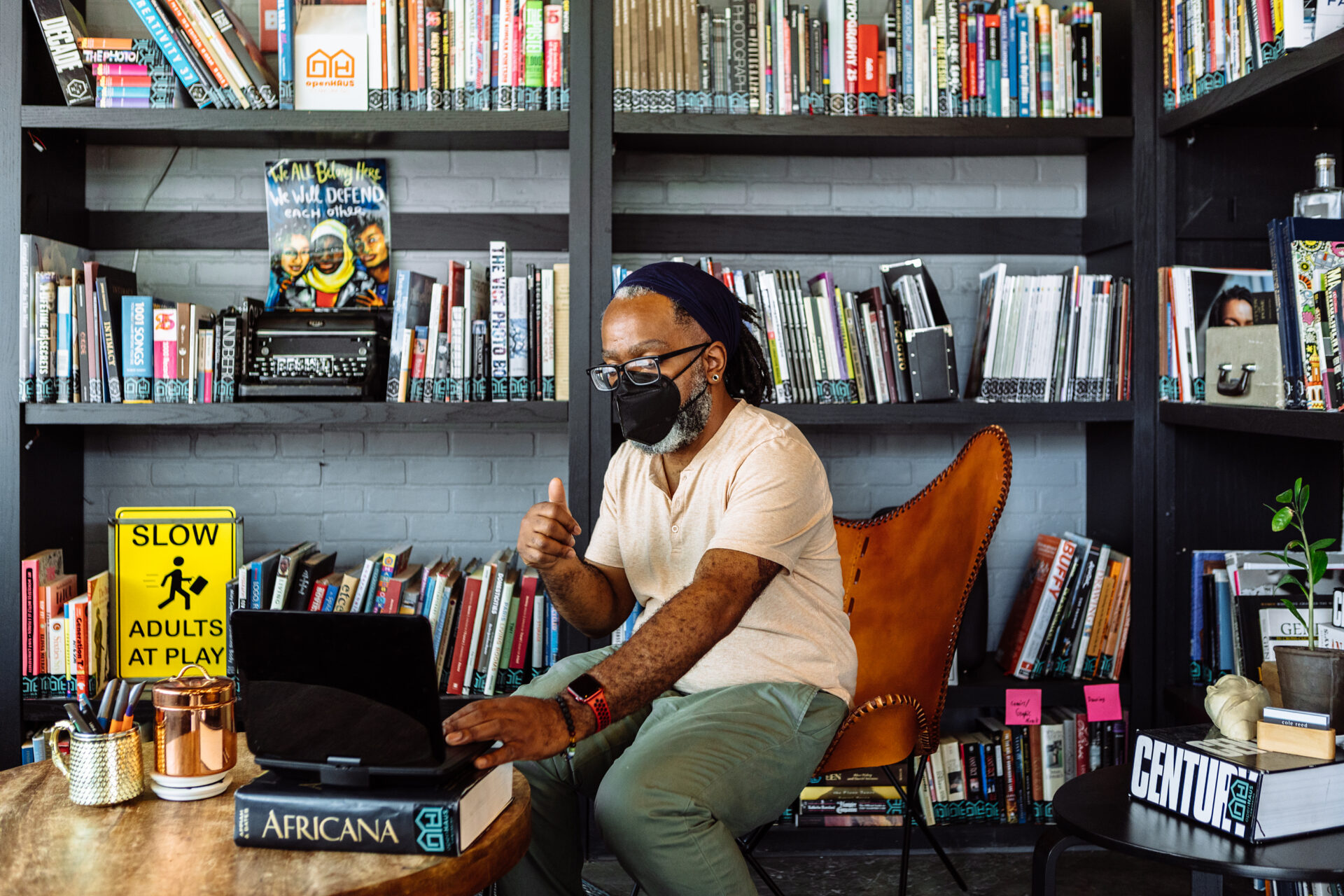 A Deaf Black man wearing glasses and a KN-95 mask adjusts a laptop with one hand while signing with the other during a video chat. The man sits at a coffee table inside a community library packed with books and posters.