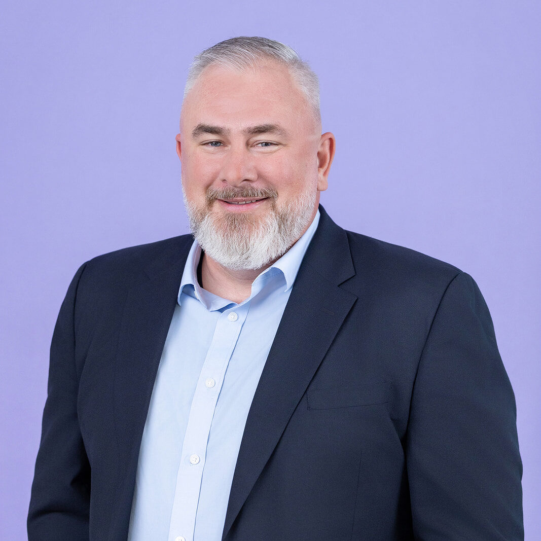 A person with short gray hair and a neatly trimmed white beard wearing a light blue collared shirt and a dark suit jacket stands in front of a light purple background.