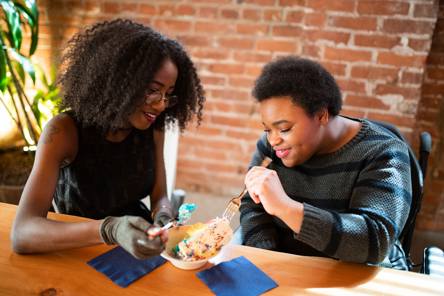 Close-up of two disabled Black people (a femme wearing compression gloves and a non-binary person in a power wheelchair) sitting next to each other and sharing a slice of cake on a date.
