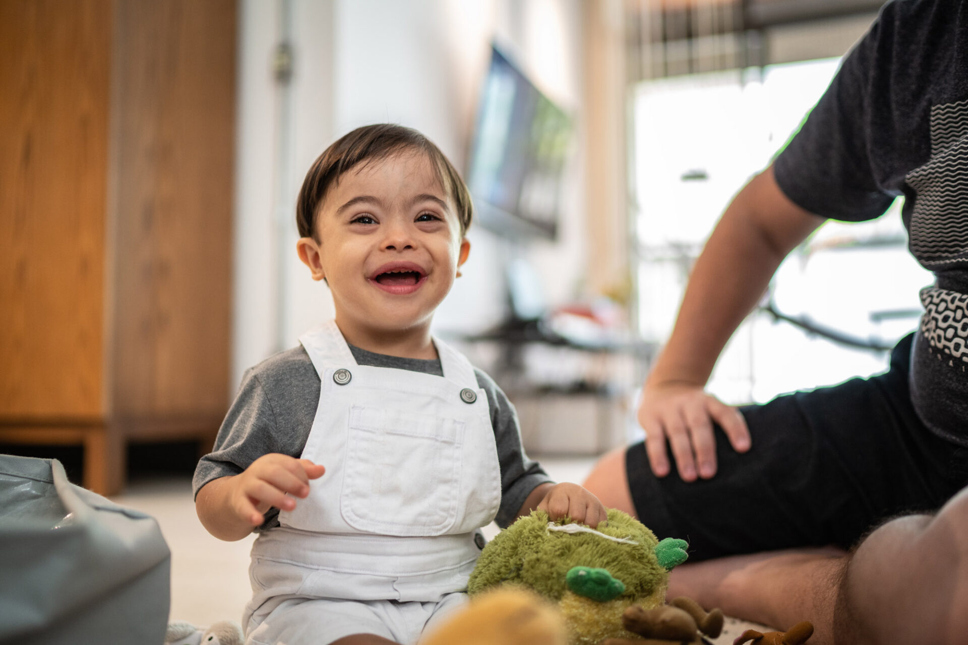 Little boy with Down Syndrome playing at home with a green stuffed animal while their caregiver sits beside them.