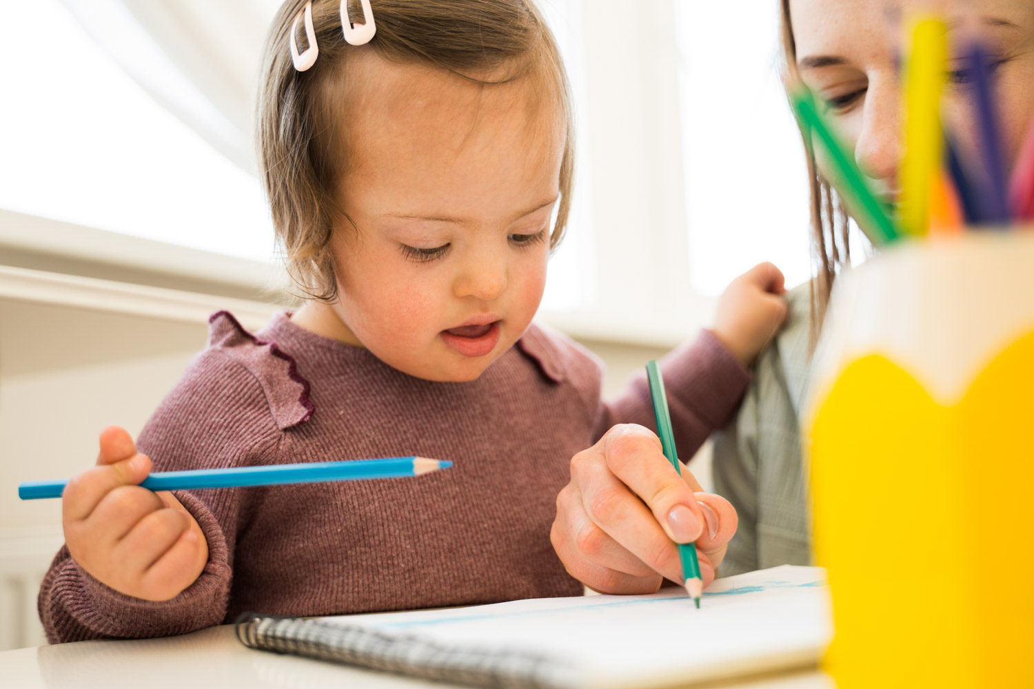 Two year old girl with Down Syndrome spending time drawing with her mother on a sheet of paper at the table.