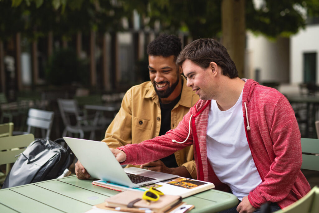 A young man with Down Syndrome with his mentoring friend sitting outdoors in cafe using laptop.
