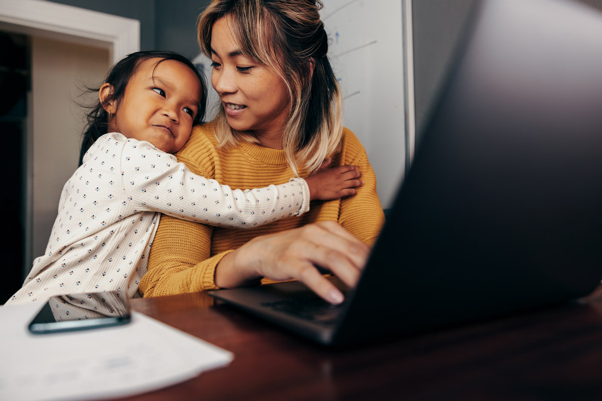 Little girl embracing her mom while she smiles back at her while typing on a black laptop.