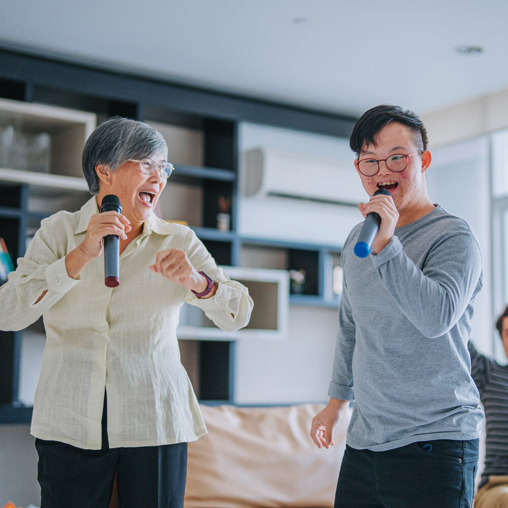Young Chinese man with Down syndrome singing karaoke with his grandmother having fun in living room.
