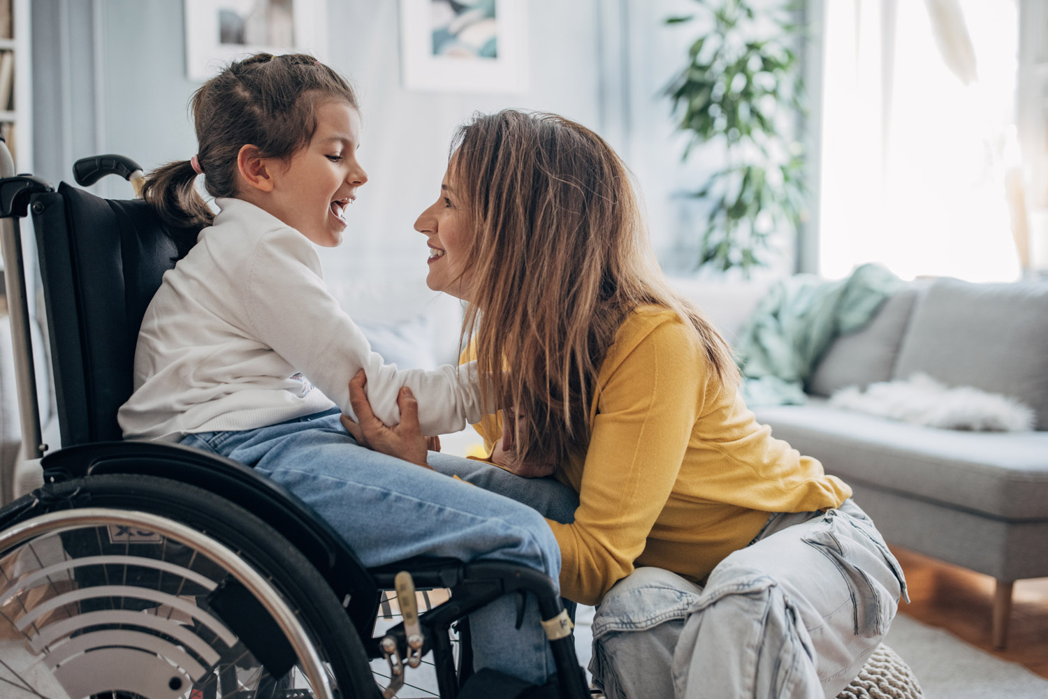 Mother and young daughter who is in wheelchair enjoying time together in living room at home.
