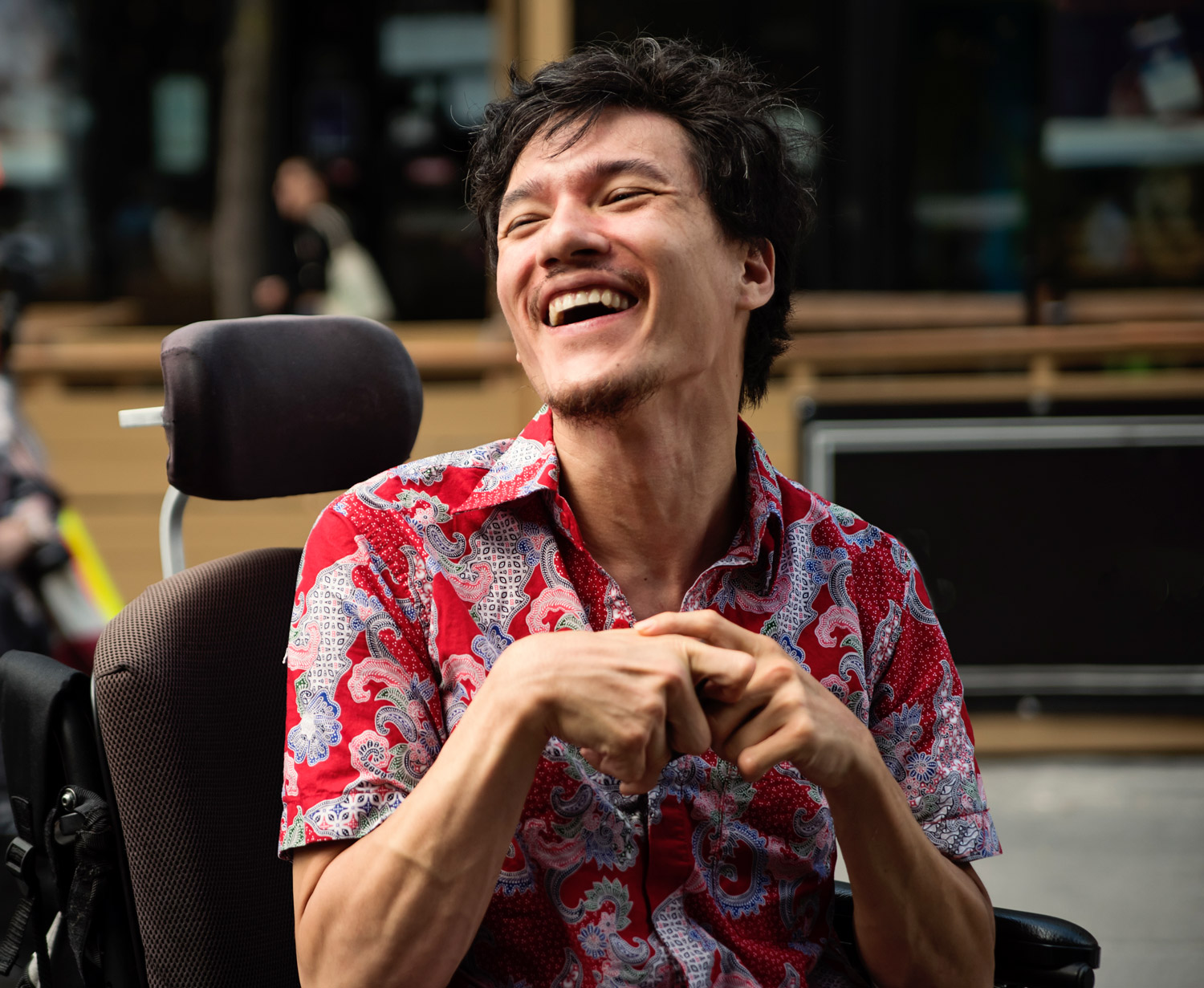 Young man with cerebral palsy in a wheelchair laughing while outdoors in city. He is of Indonesian ethnicity and a member of the LGBTQ+ community.