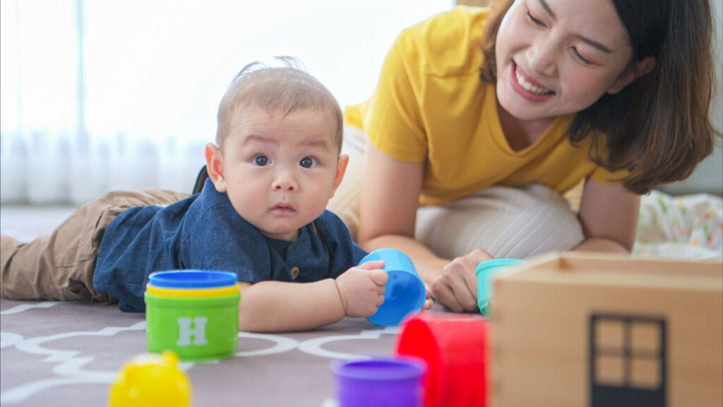 A tiny child cheerfully plays with toys on the floor of the living room with an adult woman.