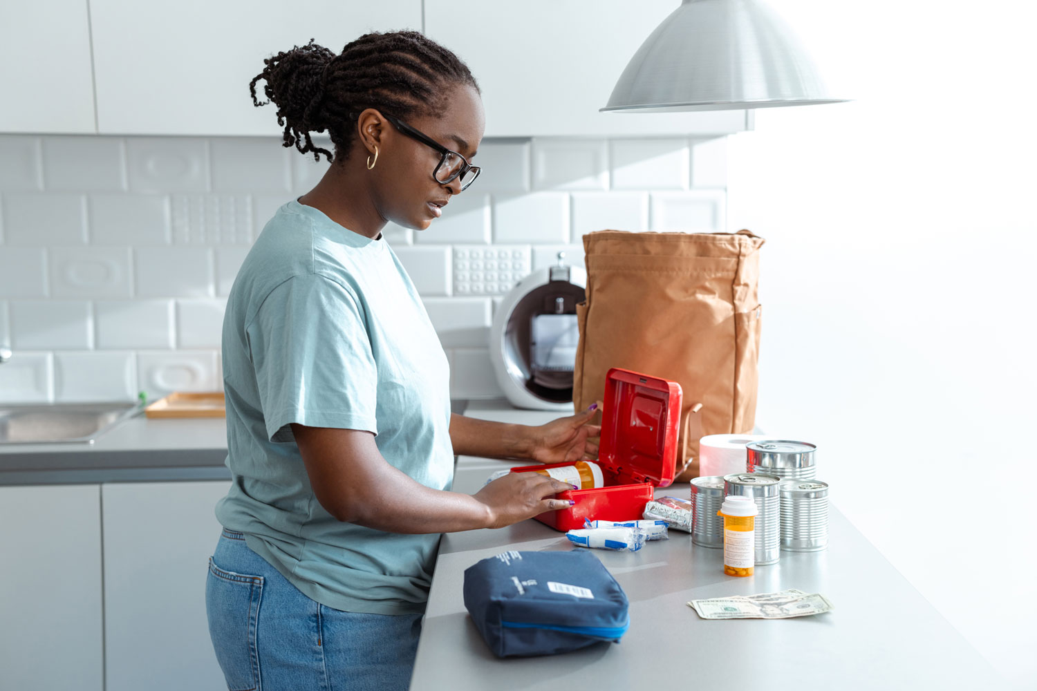A young, Black woman organizes a first aid kit at the kitchen counter, placing supplies like medications and bandages into the red medical box, with a paper bag and cans nearby.