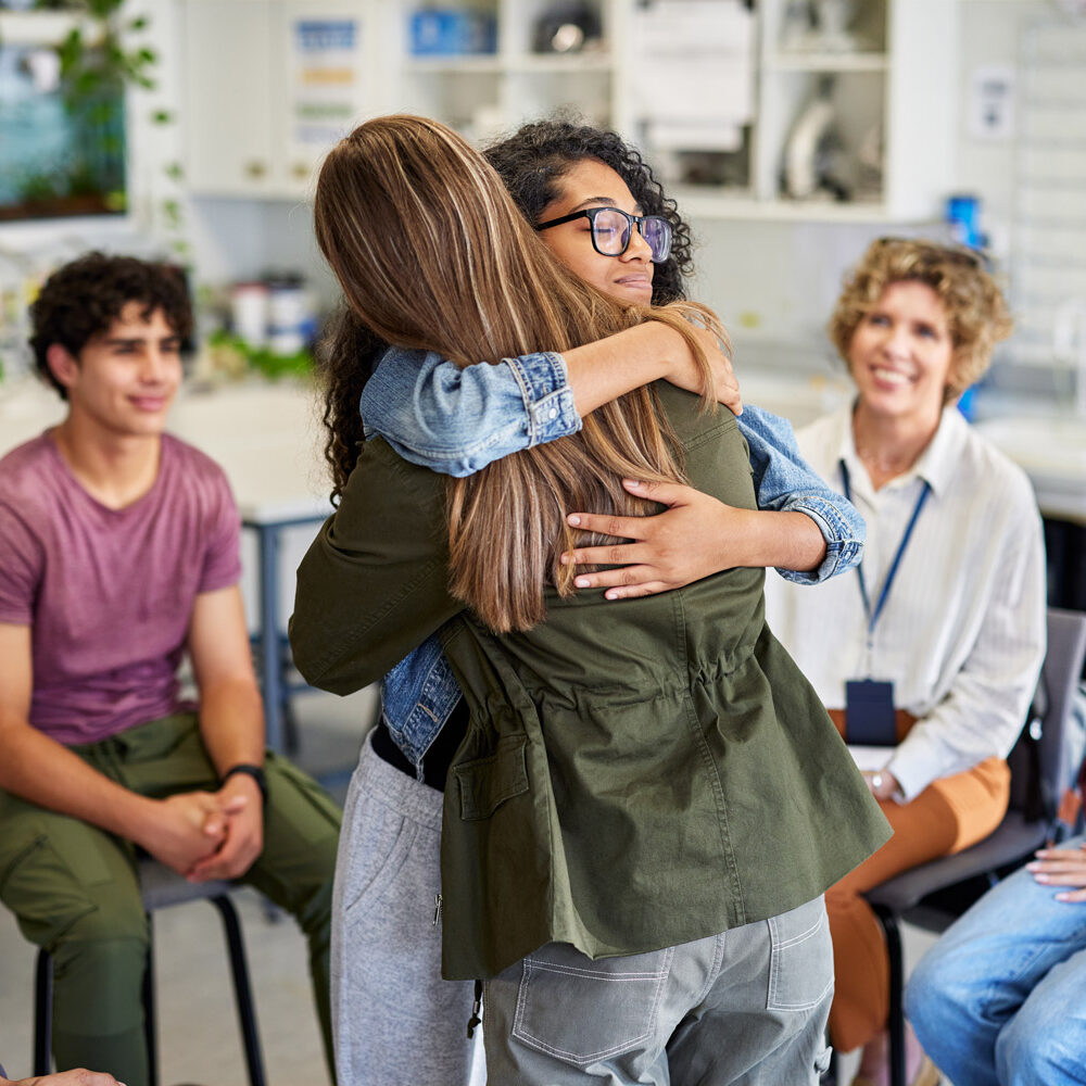 Duas alunas abraçadas durante uma sessão de aconselhamento em grupo, oferecendo conforto e ligação emocional. Colegas de turma de várias etnias observam esse momento de apoio com expressões atentas e compassivas. A terapeuta sorri ao fundo, testemunhando o resultado positivo da sessão.