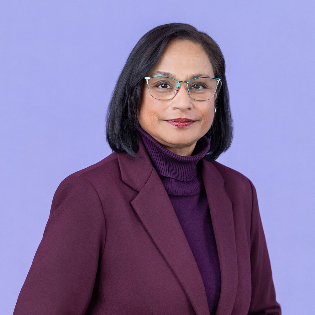 A person with short dark hair and glasses wearing a dark purple turtleneck and a maroon blazer stands in front of a light purple background.